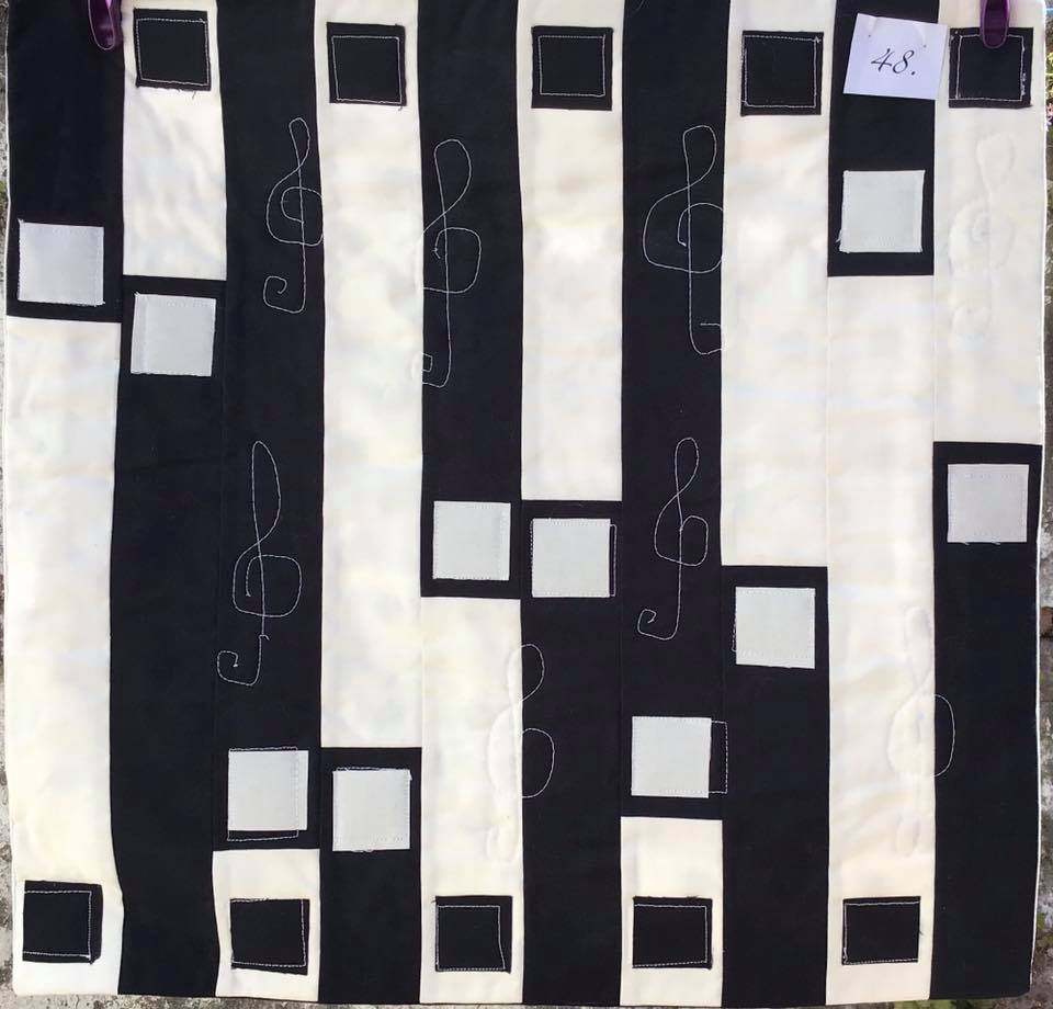 A small, square quilt with a black and white design. The quilt features vertical stripes of black and white fabric, with white treble clefs and squares scattered across the surface.