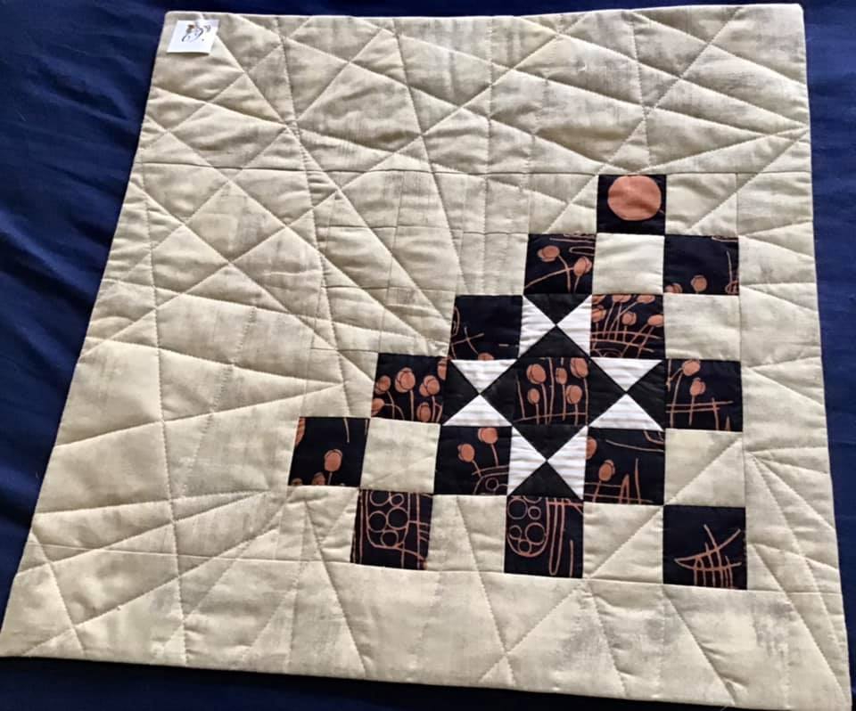 A small, square quilt with a beige background. The quilt features a pattern of black and white squares arranged in a diagonal line, with some squares incorporating a red flower design. The background is quilted with a complex pattern of intersecting lines.