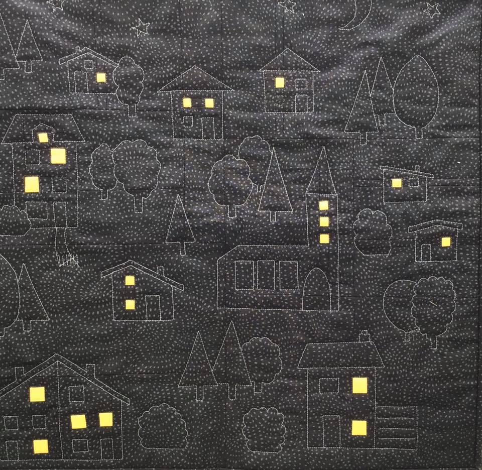 A small, square quilt with a dark background. The quilt features a whimsical night scene with white outlines of houses, trees, and a church. The houses have small, yellow squares for windows, suggesting they are lit up. The background is filled with tiny white dots.
