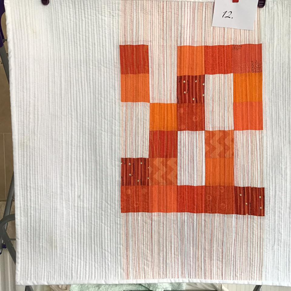 A small, square quilt with a white background. The quilt features a pattern of orange squares arranged in a vertical line on the right side of the quilt. The white background has a subtle, textured pattern.