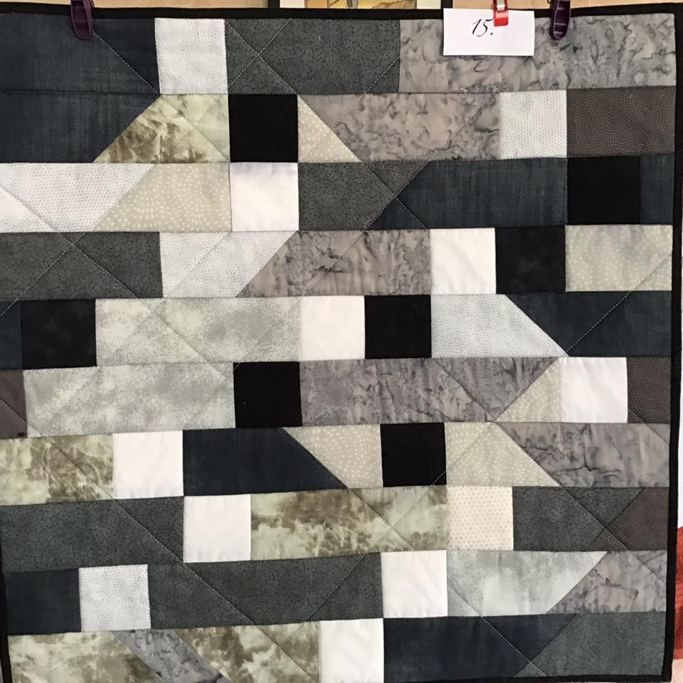 A small, square quilt with a patchwork design. It features various shades of grey and black fabric arranged in a geometric pattern of squares and triangles.