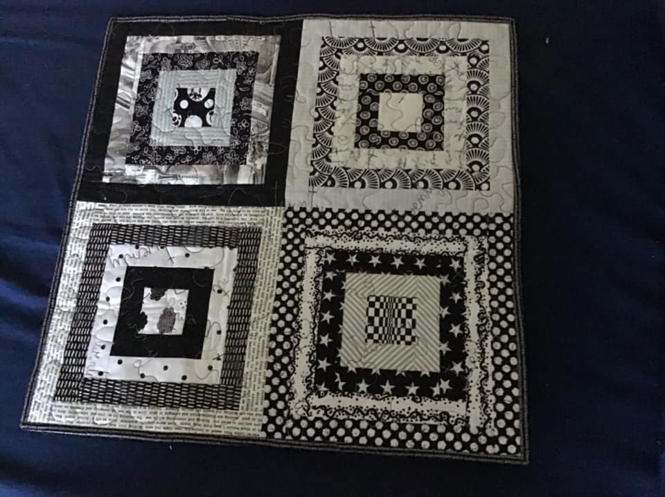 A square quilt with a black and white colour scheme. The quilt is divided into four sections, each featuring a different pattern within a square frame. The patterns include stripes, polka dots, stars, and text. The background is quilted with a subtle, linear pattern.
