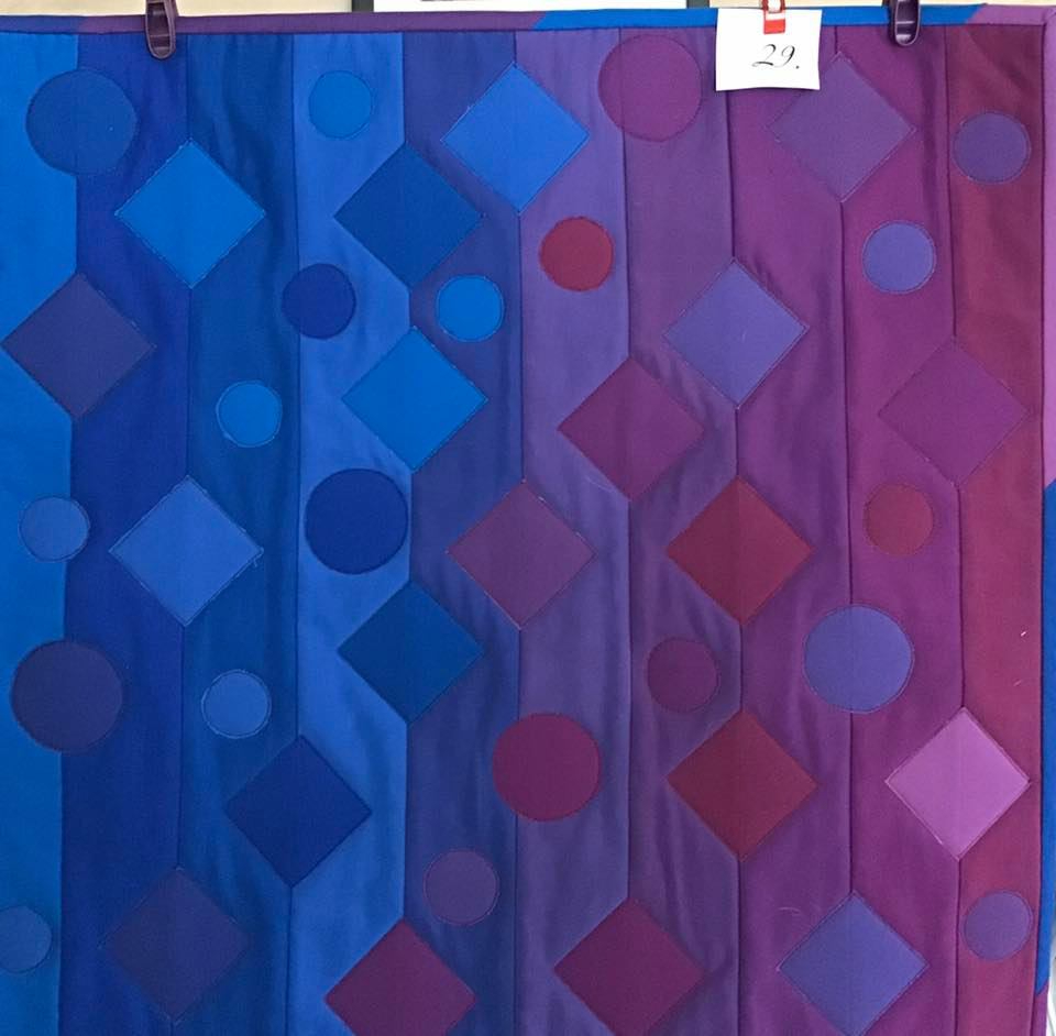 A small, square quilt with a gradient design. It features shades of blue and purple arranged in vertical stripes. There are also circles and diamonds scattered throughout the quilt.