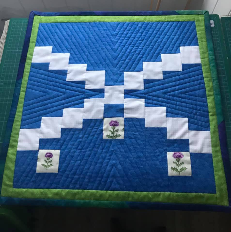 A small, square quilt with a Scottish theme. It features a blue background with a white diagonal cross, representing the Saltire flag of Scotland. There are three embroidered thistles, the national flower of Scotland, on the white cross. The quilt has a green border.