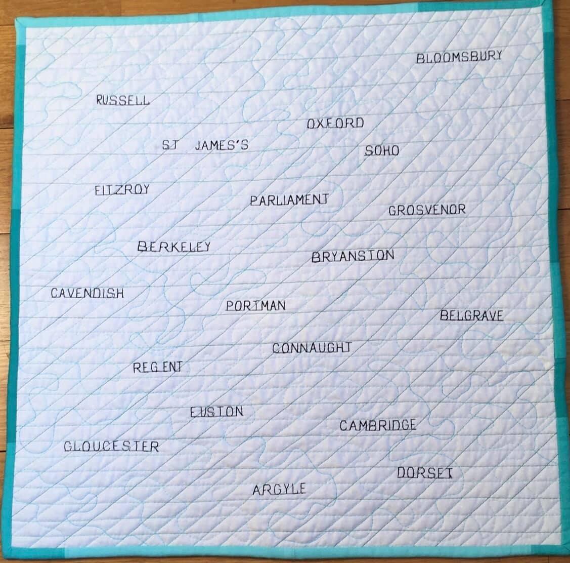 A quilt with a white background and a blue border. The quilt features the names of London streets arranged in a crossword puzzle style. The streets include Russell, St. James's, Oxford, Soho, Grosvenor, Berkeley, Portman, Connaught, Belgrave, Regent, Euston, Cambridge, Dorset, Gloucester, and Argyle.