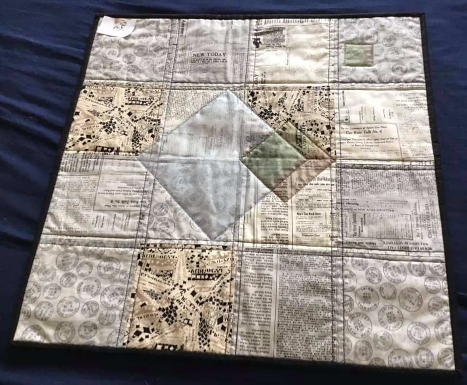 A small, square quilt with a white background and a black border. The quilt features a patchwork design with squares of fabric featuring newspaper clippings and advertisements. In the center is a diamond shape made up of smaller squares.