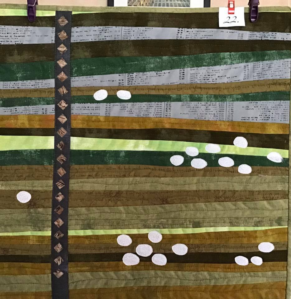 A small, rectangular quilt with a landscape-like design. It features horizontal strips of green and brown fabric, with white circles scattered across them, resembling clouds. A vertical strip of black fabric with white diamonds runs along the left side.