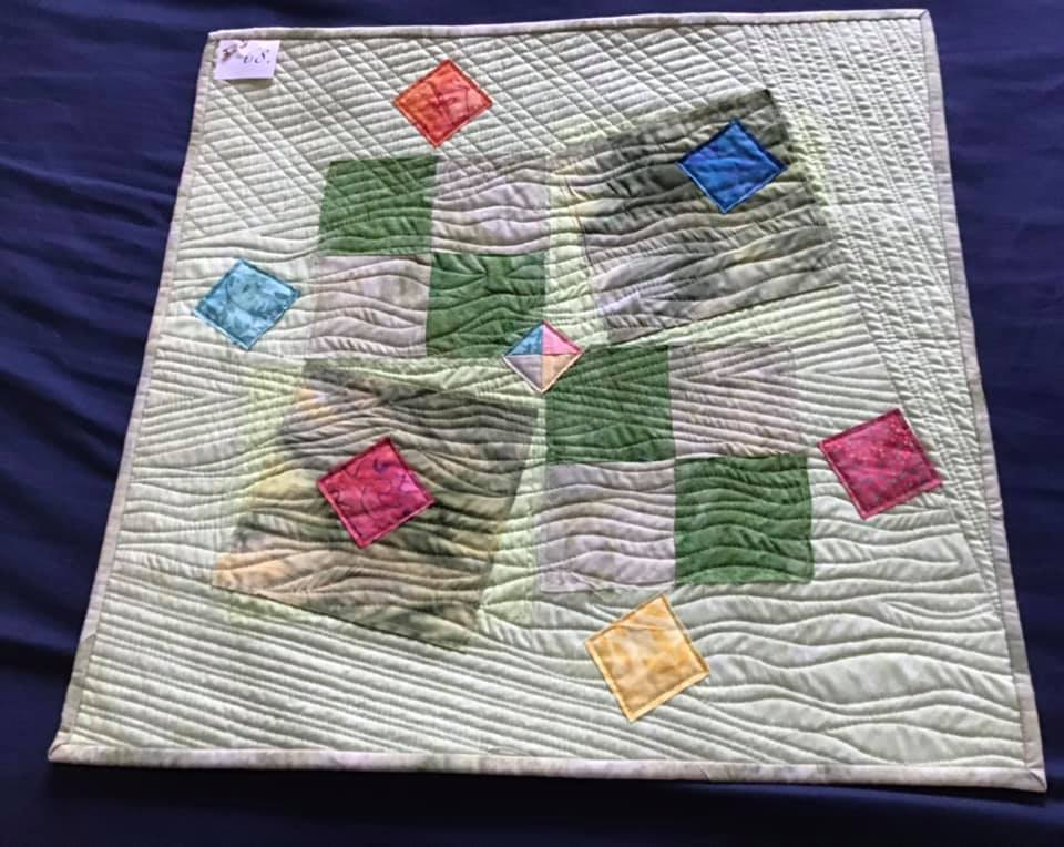 A square quilt with a green background. The quilt features a central design of overlapping squares in various colours, including green, blue, yellow, red, and pink. The background is quilted with a wavy, organic pattern.