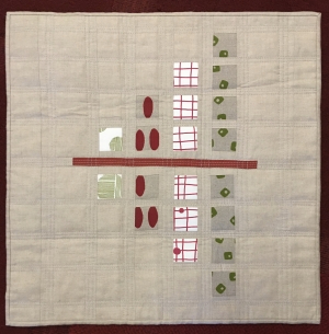 A small, square quilt with a beige background. The quilt features a symmetrical pattern of red circles, green squares, and beige shapes, arranged in rows. A red line divides the quilt horizontally.