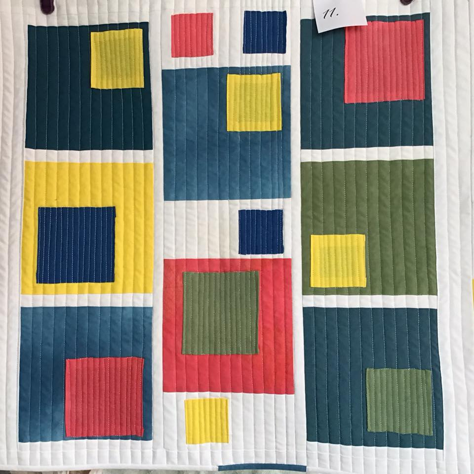 A small, rectangular quilt with a white background. The quilt features a pattern of squares in various colours, including red, yellow, green, and blue. The squares are arranged in rows and columns, creating a geometric design.