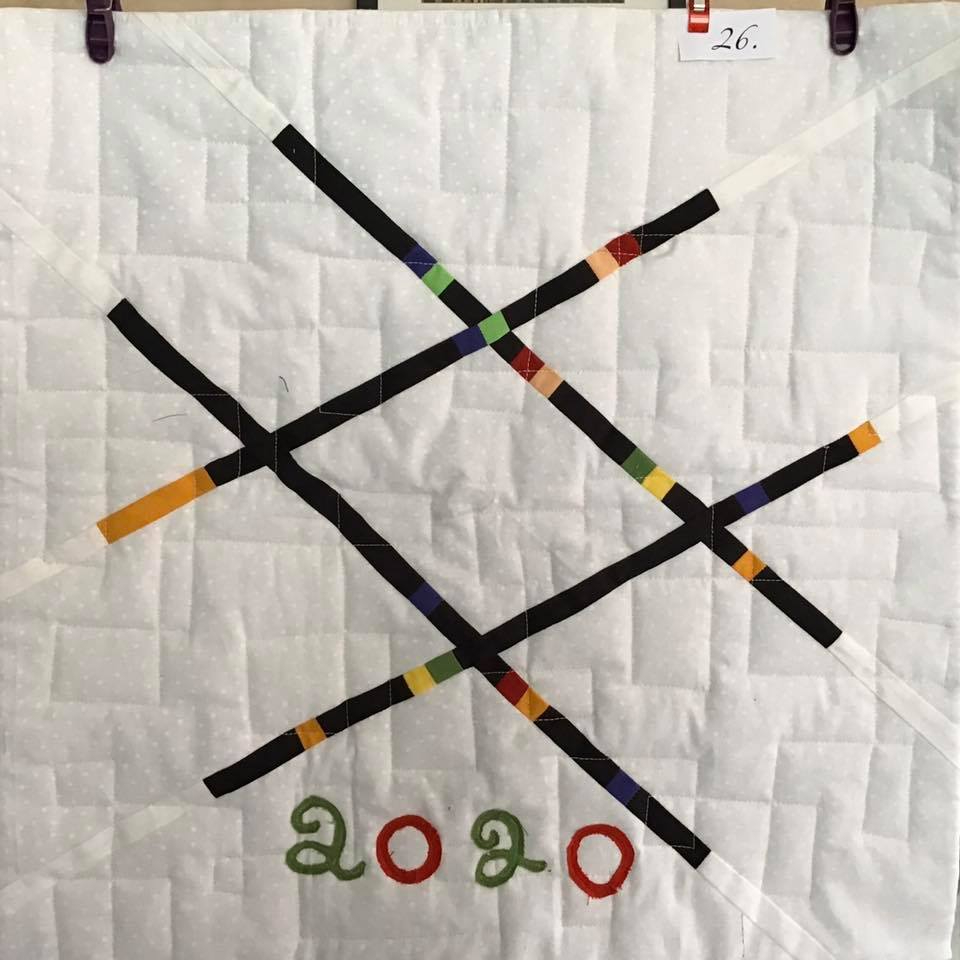 A small, square quilt with a white background. The quilt features a large, black "X" shape with colourful lines intersecting it. The year "2020" is embroidered in colourful thread at the bottom of the quilt.