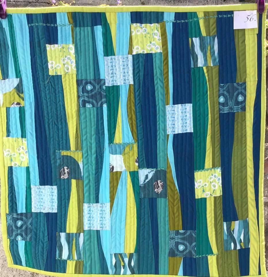 A small, square quilt with a patchwork design. It features vertical strips of fabric in shades of green and blue, with some squares and rectangles added for contrast. The quilt has a textured, quilted pattern.