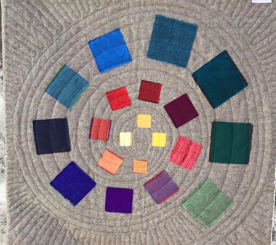 A small, square quilt with a grey background. The quilt features a circular pattern of coloured squares arranged in a spiral. The colours range from blues and purples to reds, oranges, and yellows. The background is quilted with concentric circles.