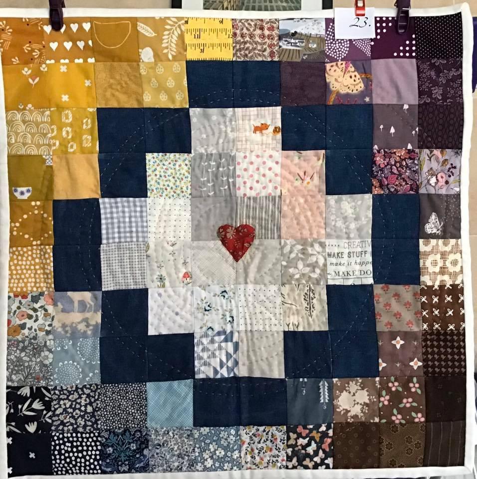 A small, square quilt with a patchwork design. It features a variety of colourful fabrics with different patterns, including hearts, flowers, and animals. The quilt is framed by a dark blue border.