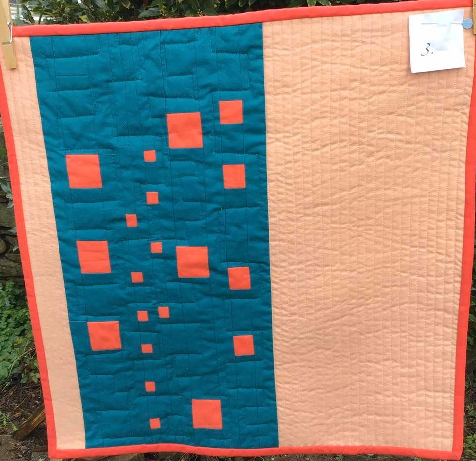 A square quilt with a two-tone design. The left side is teal with a pattern of orange squares of varying sizes. The right side is peach-coloured. The quilt has an orange border.