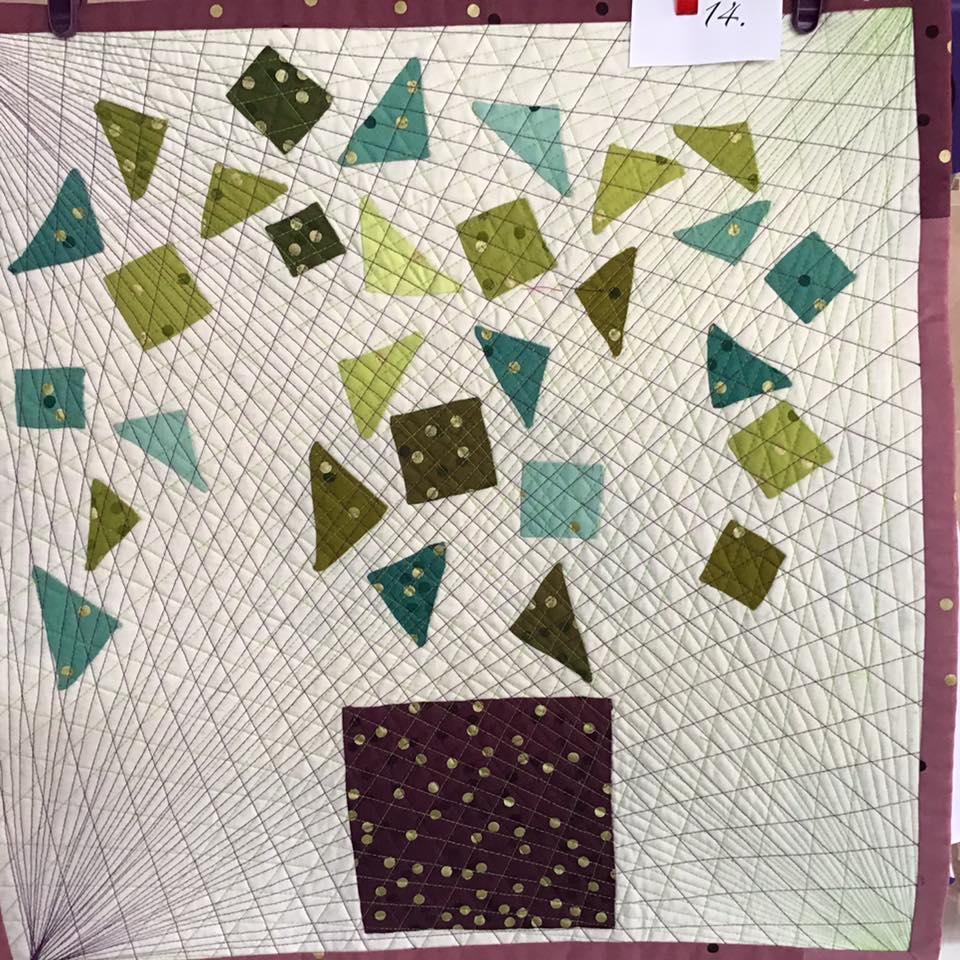 A small, square quilt with a cream-coloured background. The quilt features a design of green and blue triangles and squares arranged in a tree-like shape. The "trunk" of the tree is a brown rectangle. The background is quilted with a radiating pattern of thin lines.