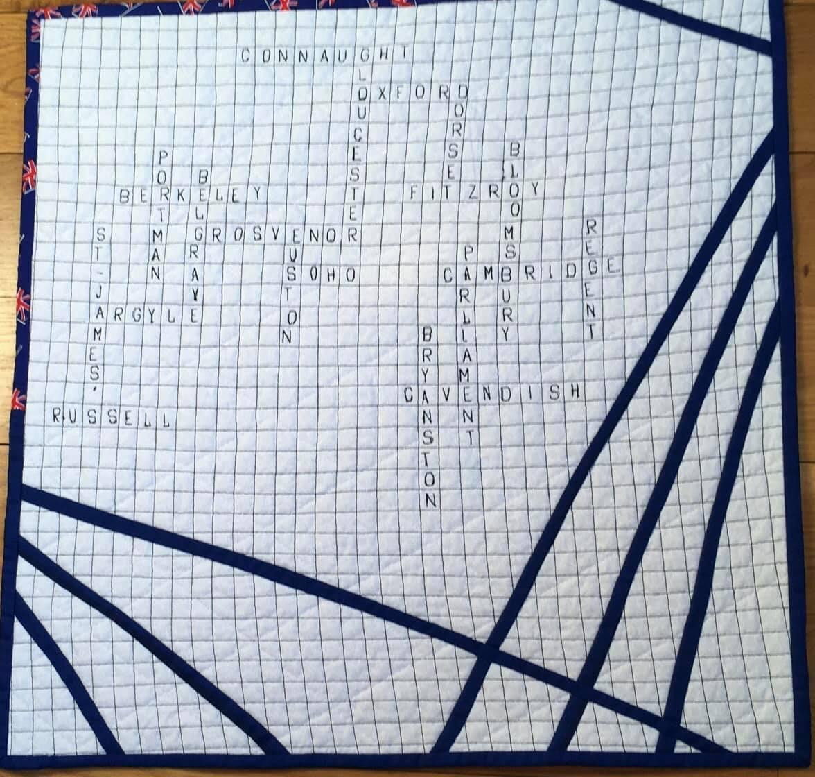 A crossword quilt with blue fabric and white gridlines. The crossword puzzle features words related to London streets, including Russell, James, Argyle, Soho, and more. The quilt has a black border with diagonal lines.