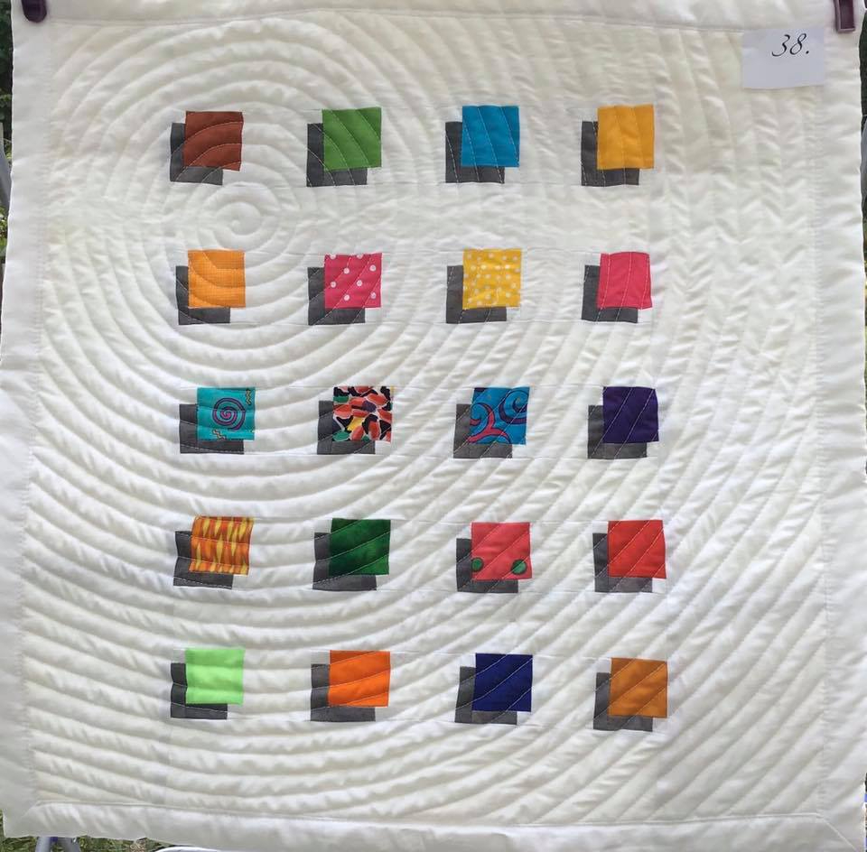 A small, square quilt with a white background. The quilt features a pattern of colourful squares arranged in a circular pattern. The background is quilted with concentric circles.