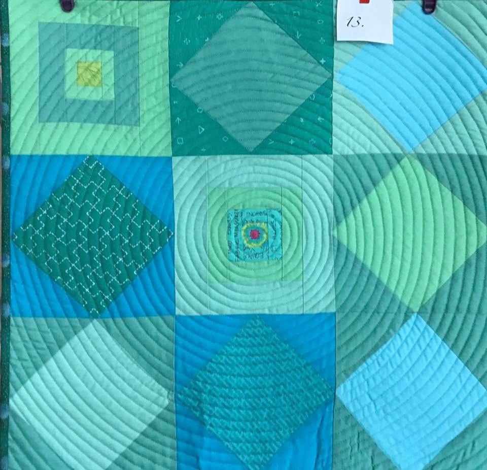 A small, square quilt with a patchwork design. It features various shades of green and blue fabric arranged in a grid pattern. The central square has a circular pattern of concentric rings, while the other squares have different geometric shapes and patterns.