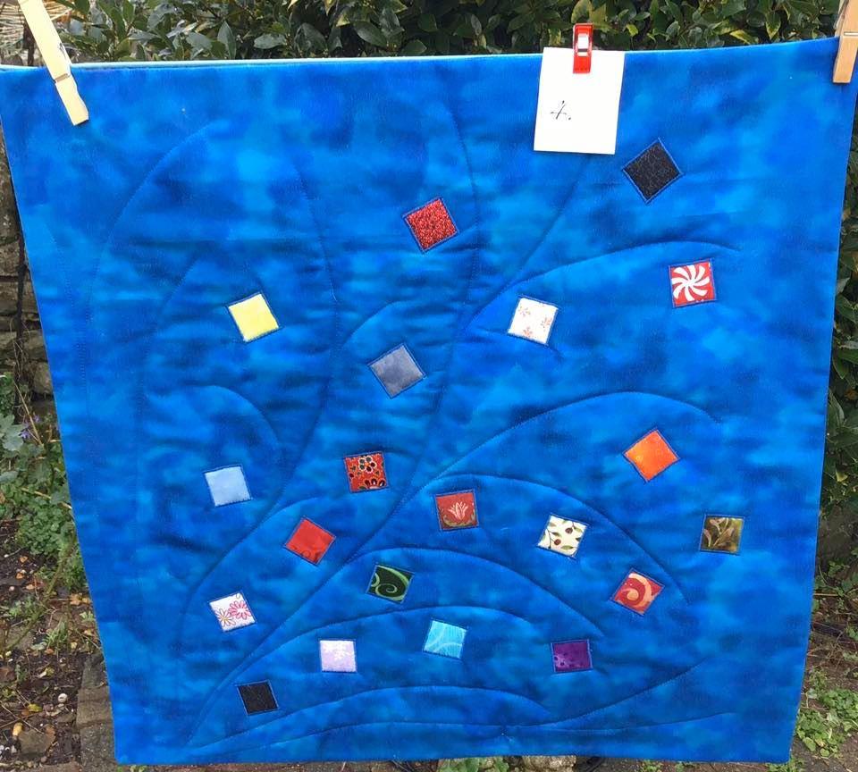 A square quilt with a vibrant blue background. The quilt features a pattern of curved lines radiating outwards from the centre, creating a flower-like shape. There are various coloured squares and rectangles scattered across the blue background.