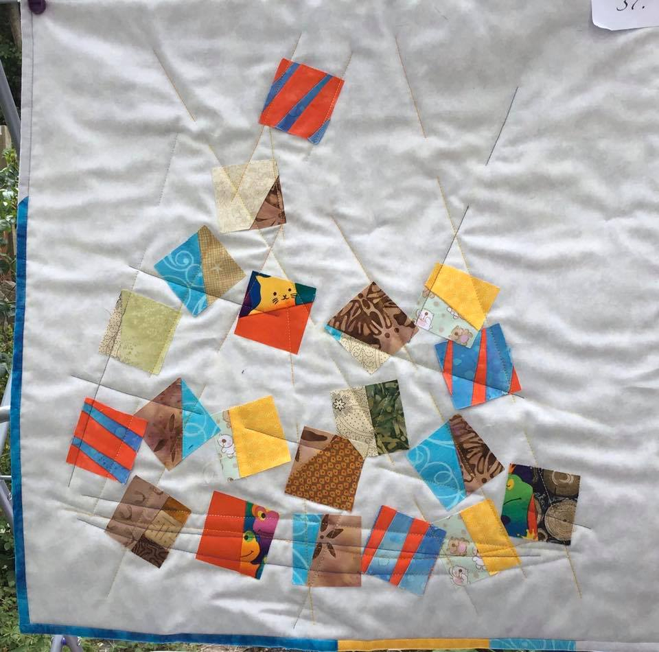 A small, square quilt with a white background. The quilt features a playful pattern of colourful squares and triangles arranged in a seemingly random fashion. Some of the squares have patterns or designs on them.