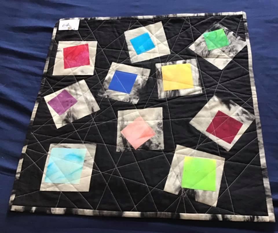 A small, square quilt with a black background. The quilt features colourful squares of various sizes, each with a unique pattern or texture. The squares are scattered across the black background, which is quilted with a geometric pattern.