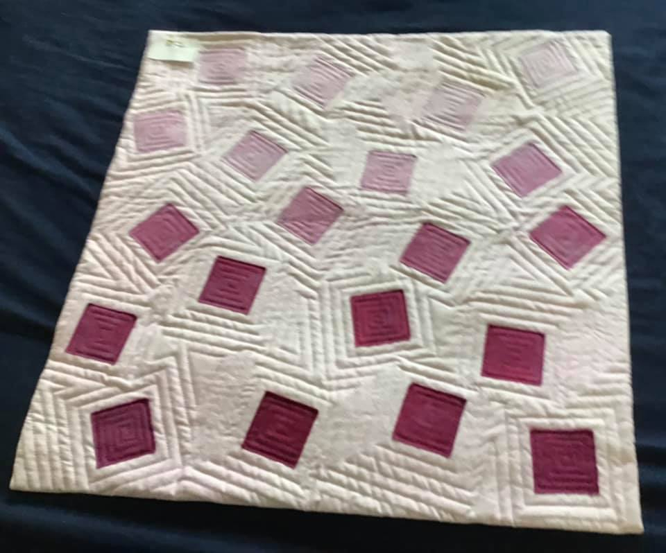 A small, square quilt with a white background. The quilt features a pattern of pink squares of various sizes, arranged in a seemingly random fashion. The squares are made of fabric in different shades of pink, creating a gradient effect. The background is quilted with a geometric pattern of lines and squares.