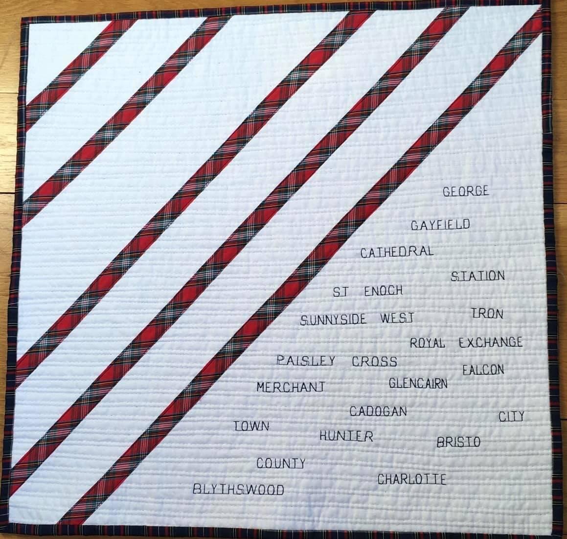 A quilt with a white background and red, diagonal stripes. The names of Glasgow streets are embroidered on the white space between the stripes, including George, Gayfield, Cathedral, St. Enoch, Station, Sunnyside, West, Royal Exchange, Paisley Cross, Merchant, Town, Hunter, Cadogan, Bristol City, County, Blythswood, and Charlotte.