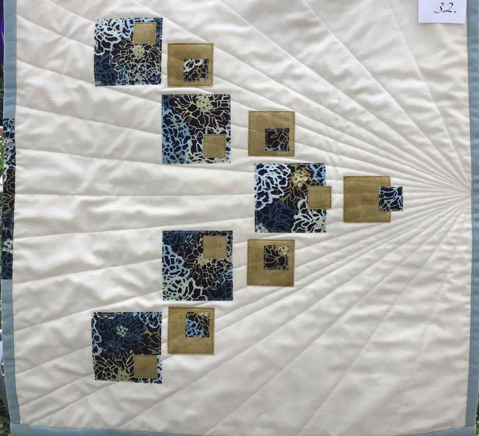 A small, square quilt with a white background. The quilt features a pattern of squares in blue and brown, arranged in a diagonal line. The squares are made of fabric with floral patterns. The background is quilted with radiating lines.