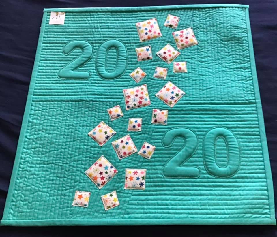 A small, square quilt with a turquoise background. The quilt features the number "2020" written twice in large, white, quilted letters. Between the numbers, there is a trail of colourful, quilted squares with star patterns.