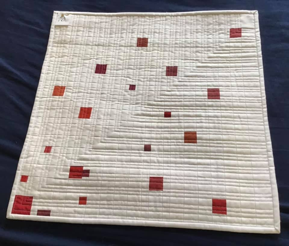 A small, square quilt with a white background. The quilt features a pattern of red squares of various sizes, arranged in a diagonal line. The background is quilted with a grid-like pattern.