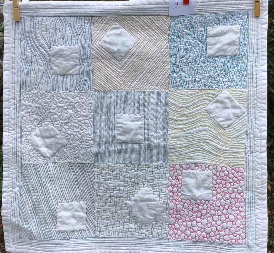 A small, square quilt with a white background. The quilt is divided into nine sections, each with a different pattern of stitching, including lines, dots, and geometric shapes.
