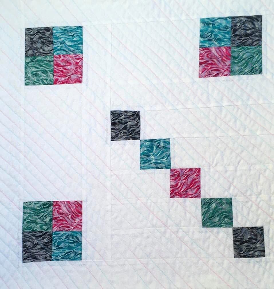 A small, square quilt with a white background. The quilt features a pattern of colourful squares arranged in a diagonal line. The squares are made of fabric with a wavy, marbled pattern in shades of pink, blue, and gray. The background is quilted with a subtle, diagonal line pattern.