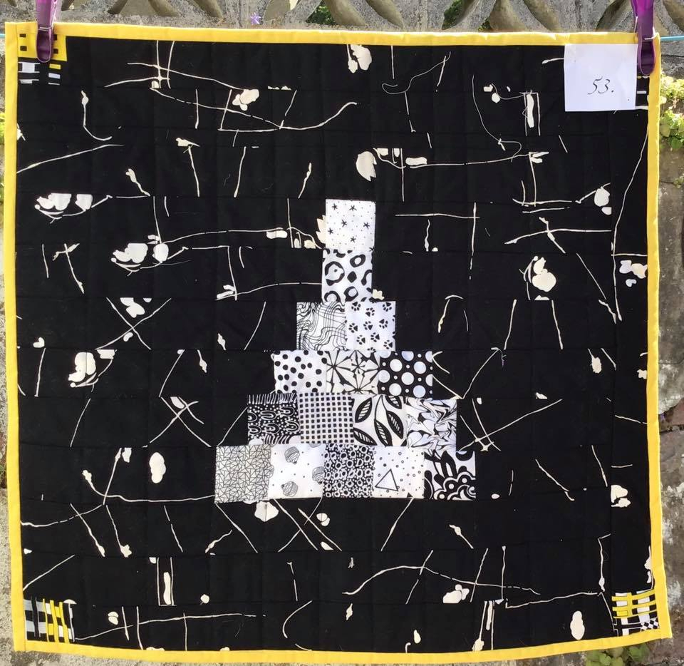 A small, square quilt with a black background. The quilt features a white, pyramid-shaped design made up of smaller squares with various black and white patterns. The background is quilted with a scribbled, abstract pattern.