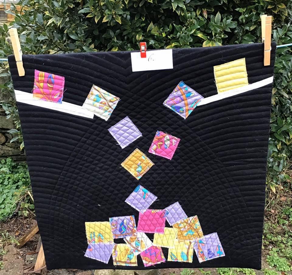 A small, square quilt with a black background. The quilt features a pattern of colourful squares arranged in a cascading design, resembling falling dominoes. The background is quilted with curved lines.
