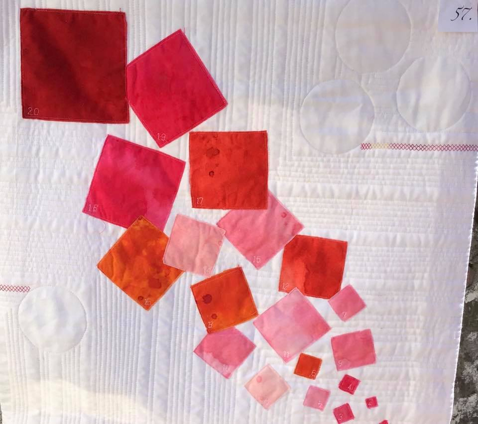 A small, square quilt with a white background. The quilt features a pattern of red squares of varying sizes, arranged in a diagonal line. Some of the squares have numbers written on them. The background is quilted with a pattern of circles and lines.
