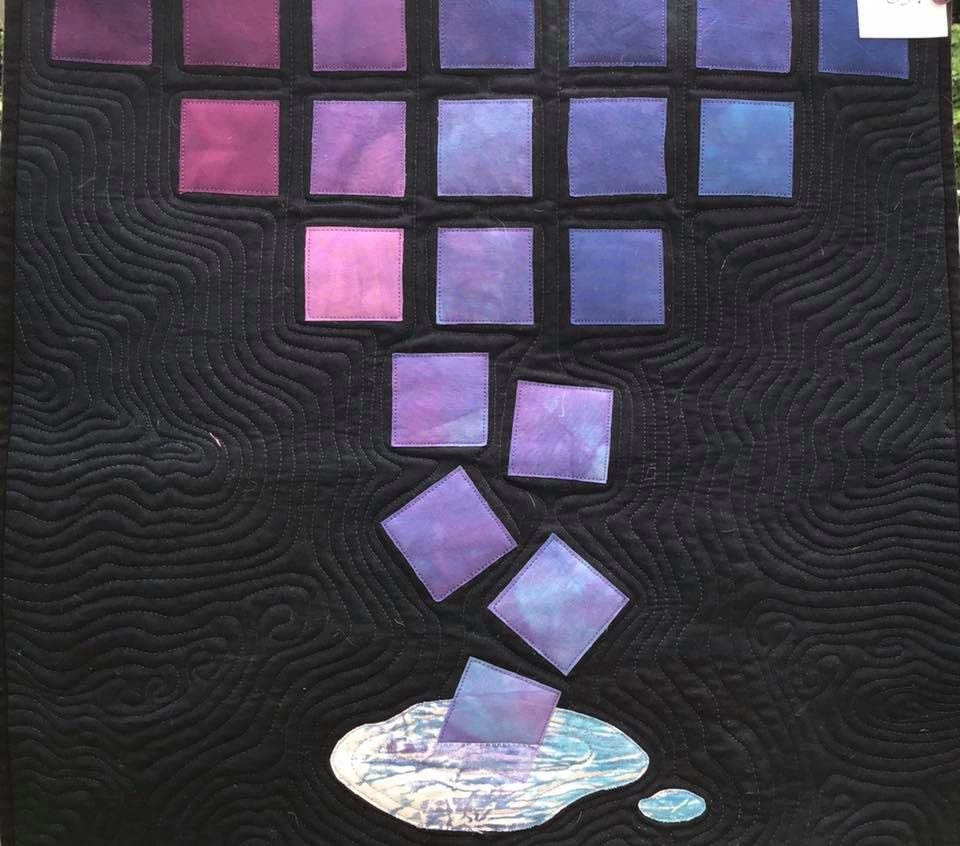 A small, square quilt with a dark background. The quilt features a pattern of purple and blue squares arranged in a cascading waterfall-like formation, with the bottom square melting into a puddle. The background is quilted with a wavy pattern.