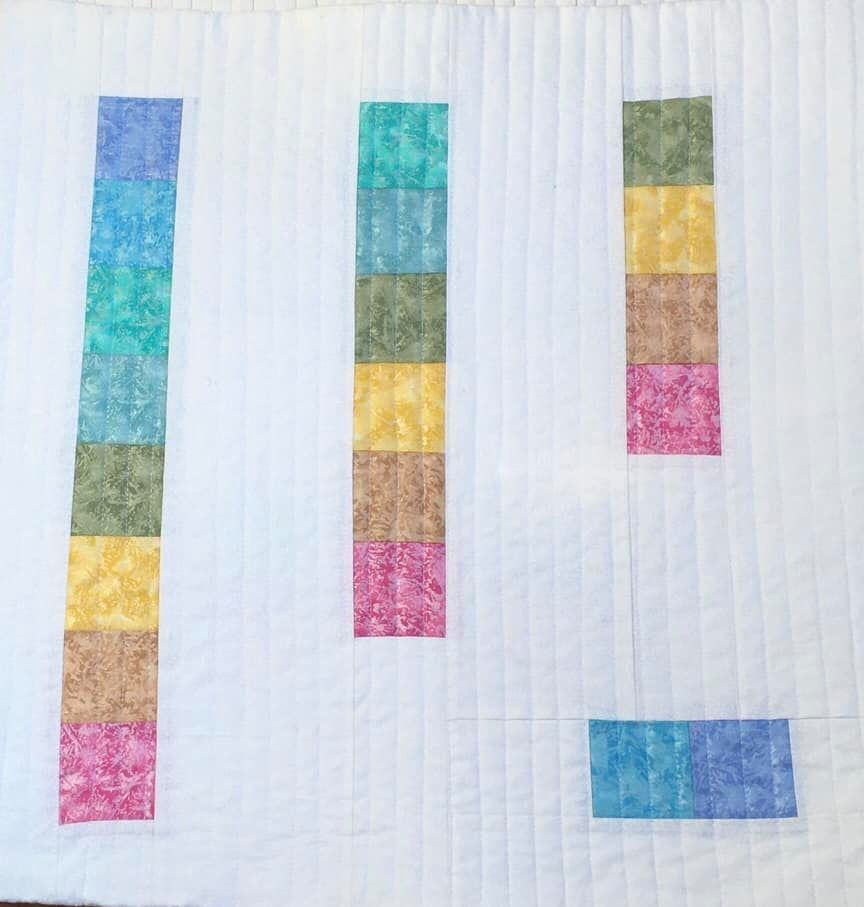 A quilt with a white background. The quilt features three vertical columns of colourful squares, each column using a different colour palette. The colours include shades of blue, green, yellow, and pink. The background is quilted with vertical lines.