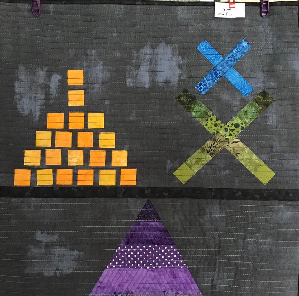 A small, square quilt with a dark grey background. The quilt features a balancing scale design. On one side is a pyramid of orange squares, and on the other side is a purple triangle made up of smaller triangles. There is also a green "X" shape above the scale.
