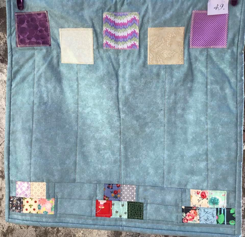 A small, square quilt with a blue background. The quilt features a patchwork design with colourful squares of various patterns and sizes, arranged in a top and bottom border.