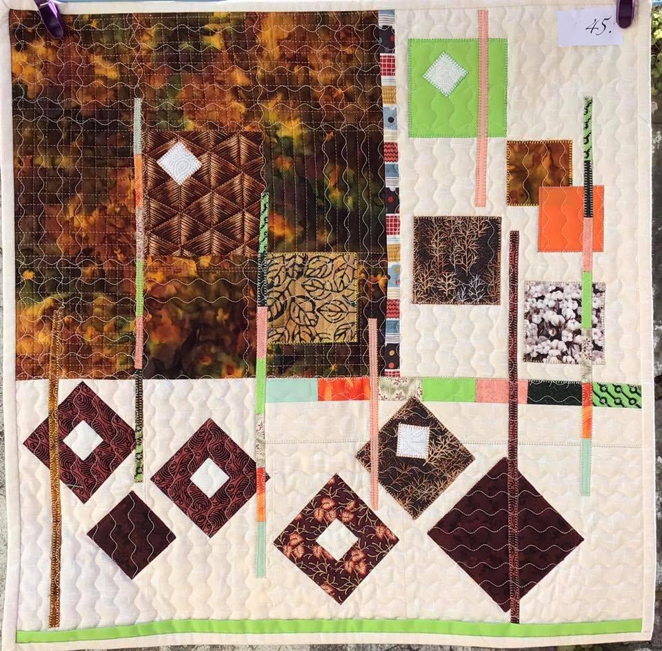 A small, square quilt with a white background. The quilt features a colourful and abstract design with squares, rectangles, and diamond shapes in various colours and patterns. The background fabric has a subtle, textured pattern.