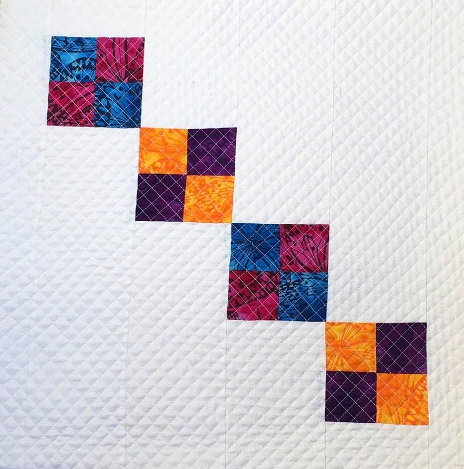 A small, square quilt with a white background. The quilt features a diagonal line of colourful squares arranged in a checkerboard pattern. The squares are made of fabric in shades of purple, orange, and blue. The background is quilted with a diamond pattern.