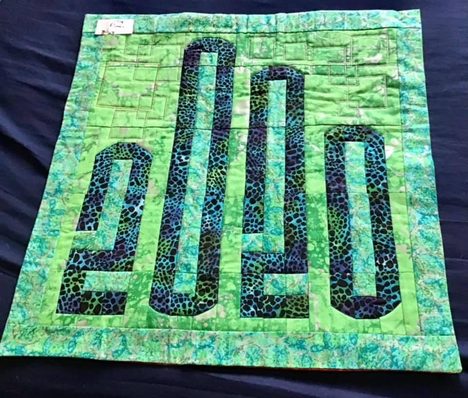 A small, square quilt with a green background and a green border. The quilt features the year "2020" written in large, black letters. The letters are filled with a pattern of blue and green shapes, resembling animal print.