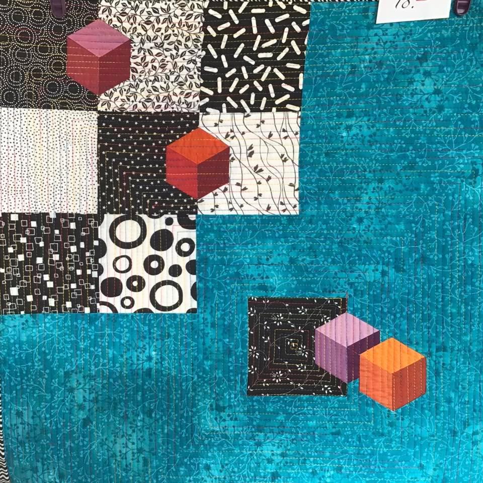 A small, square quilt with a teal background. The quilt features a patchwork design with squares of various patterns, including polka dots, stripes, and geometric shapes. There are also three-dimensional-looking cubes in pink, orange, and purple, adding depth to the design.