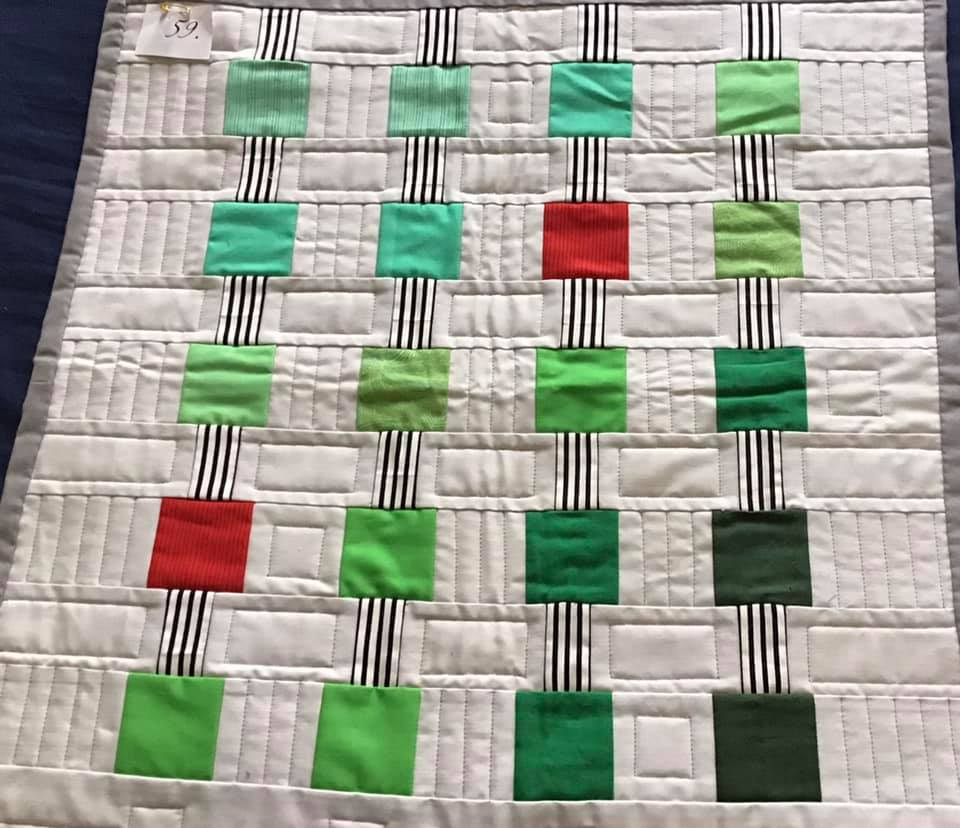 A square quilt with a white background. The quilt features a grid of colourful squares in shades of green and red. The squares are separated by black and white striped vertical lines. The background is quilted with a subtle, linear pattern.