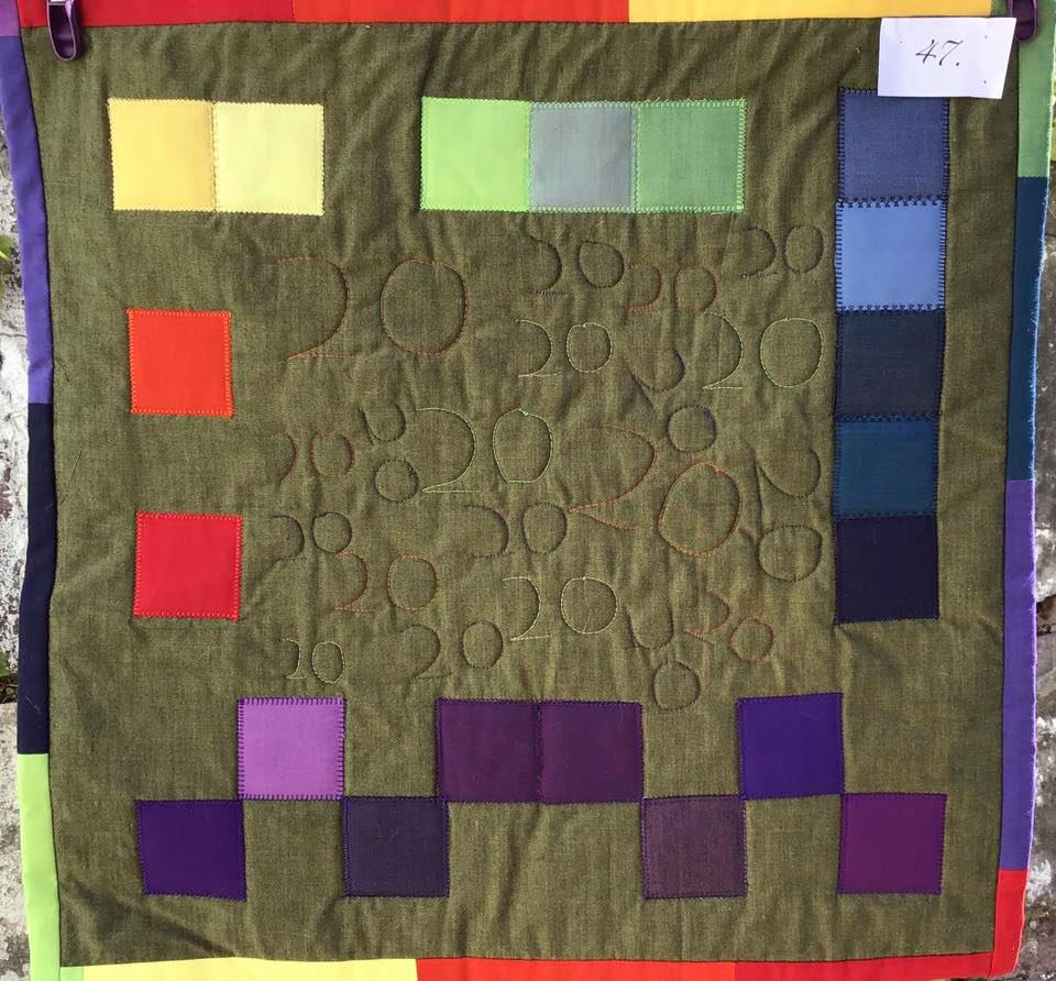 A small, square quilt with a green background. The quilt features rows of colourful squares in shades of yellow, orange, red, purple, and blue. The background is quilted with a pattern of overlapping circles.