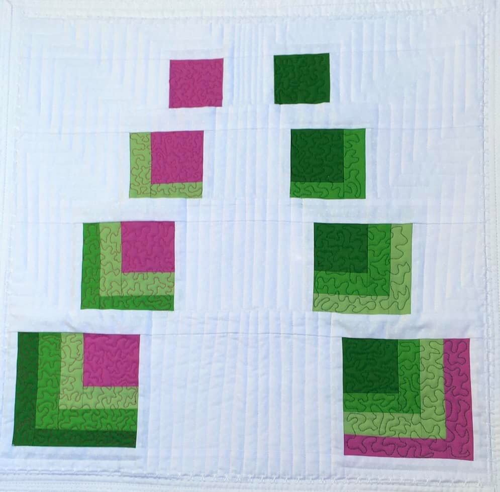 A small, rectangular quilt with a white background. The quilt features two rows of squares, one in shades of pink and the other in shades of green. The squares are arranged in a pyramid shape, with the largest squares at the bottom and the smallest at the top. The background is quilted with vertical lines.