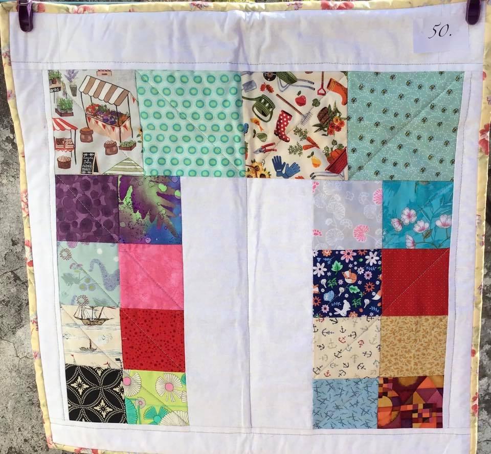A small, square quilt with a white background. The quilt features a patchwork design with colourful squares of various patterns, including floral prints, geometric shapes, and scenes like a market stall and a garden.