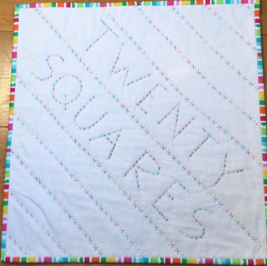 A small, rectangular quilt with a white background and a colourful border. The word "TWENTY SQUARES" is spelled out in large, colourful letters across the quilt, with a pattern of small "X" shapes in between the letters.