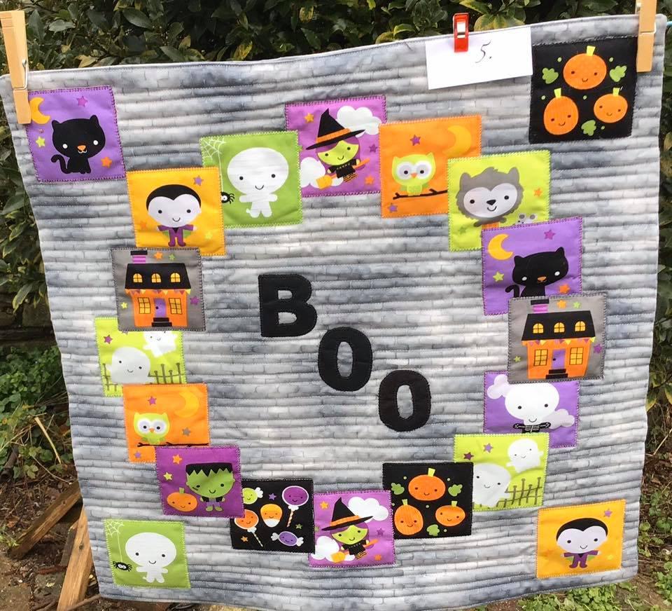 A small, square quilt with a Halloween theme. It features a grey background with a circular pattern of colourful squares depicting various Halloween characters and objects, including ghosts, pumpkins, witches, and a vampire. The word "BOO" is written in black letters in the center of the circle.